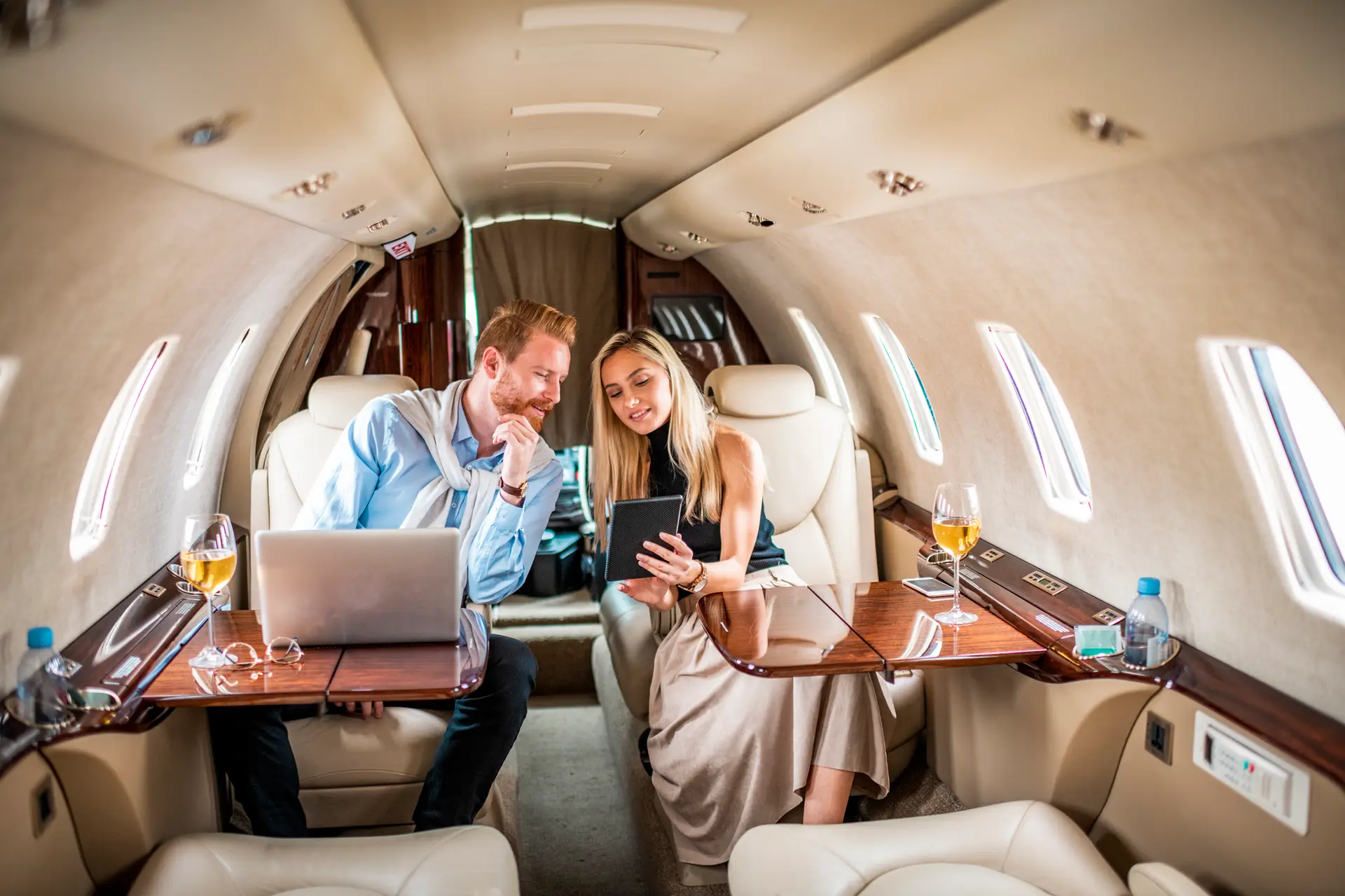Young rich couple working together while sitting in a private airplane. There is a laptop on the table in front of the man. They are looking at a digital tablet held by a woman.